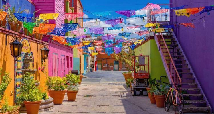 Des banderoles colorées en papel picado sont suspendues au-dessus d'une ruelle étroite bordée de bâtiments de style mexicain aux couleurs vives et de plantes en pot.