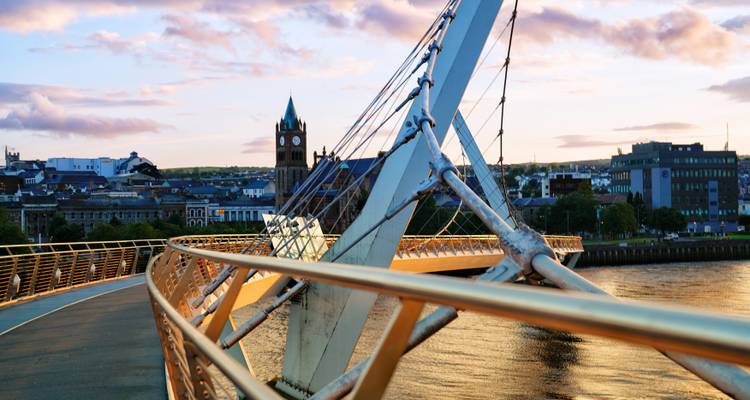 Moderne voetgangershangbrug die boven een rivier uitbolt bij zonsondergang met stadhuis klokketoren erachter.