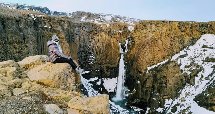 Voyageur assis sur une corniche rocheuse surplombant une haute cascade se précipitant entre des falaises saupoudrées de neige.