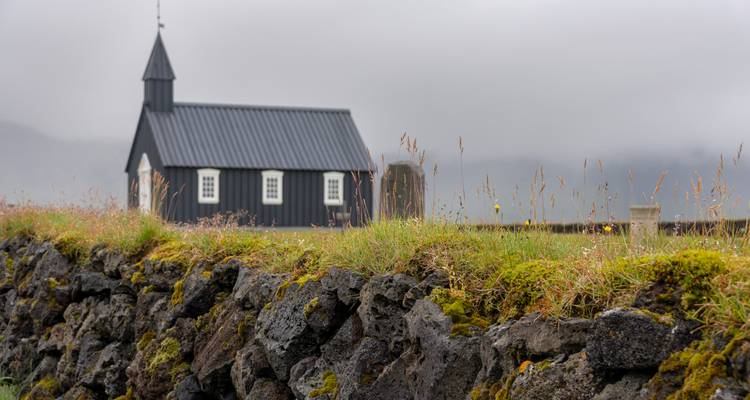 Petite église en bois noir située sur un mur de lave recouvert de mousse, sur fond de ciel gris brumeux.