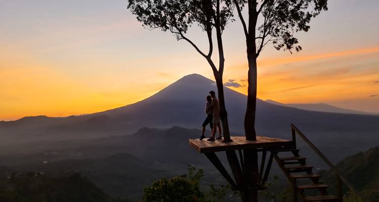 Silhouet van een stel dat elkaar omhelst op een boomplatform tegen een vurige zonsondergang en de Mount Agung.