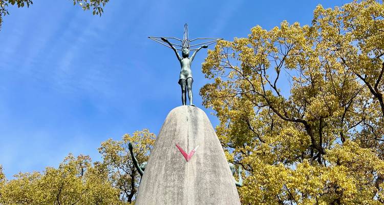 Opwaarts zicht op het standbeeld van het Kindervredes Monument met een papieren kraan in de hand tegen een levendige blauwe lucht, ingelijst door bladrijke bomen.