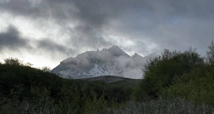 Un pic montagneux robuste et strié de neige s'élève au-dessus d'une forêt vert foncé sous des nuages brumeux épais dans une lumière tamisée.