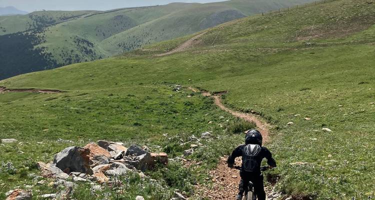 Un cycliste vêtu de noir descend un étroit sentier rocheux à travers des terres hautes verdoyantes et dégagées, parsemées de collines ondulantes.
