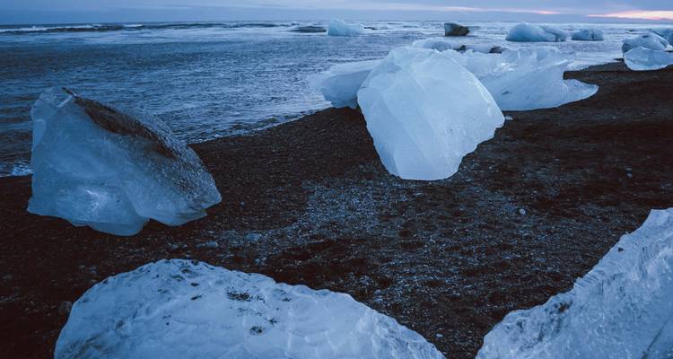 De grands blocs de glace bleue dispersés sur un rivage de sable noir au crépuscule.
