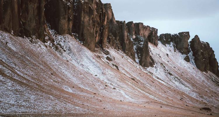 Versant montagneux abrupt et aride avec une légère couche de neige sous un ciel gris.