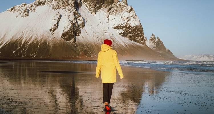 Personne en veste jaune marchant sur le sable noir mouillé avec le Vestrahorn reflété.