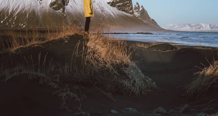Vue partielle d'une personne se tenant debout sur les dunes noires avec le Vestrahorn en arrière-plan.