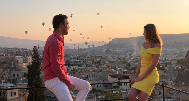 Un couple est assis sur la balustrade d'un toit en train de discuter tandis que des montgolfières se lèvent au-dessus de Goreme en contrebas au lever du soleil.