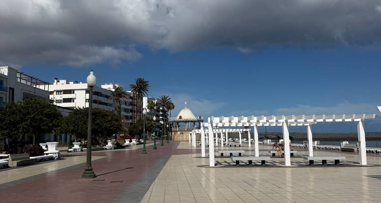Large promenade piétonne à Arrecife bordée de palmiers et de structures modernes sous un ciel nuageux mélangé