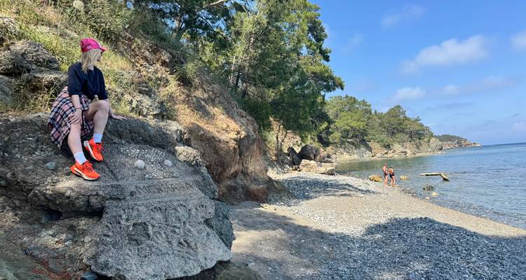 Femme sur une côte rocheuse tandis qu'une autre personne explore une plage de galets bordée de pins