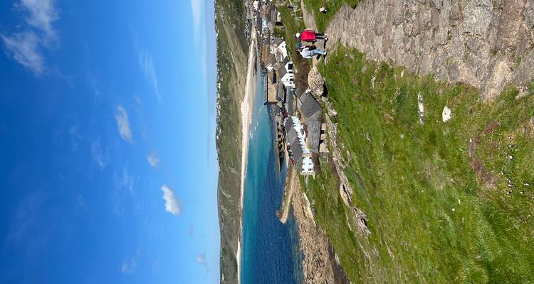 Pintoresco pueblo costero con bahía turquesa, casitas blancas, y visitantes caminando por un sendero de piedra en un día soleado.