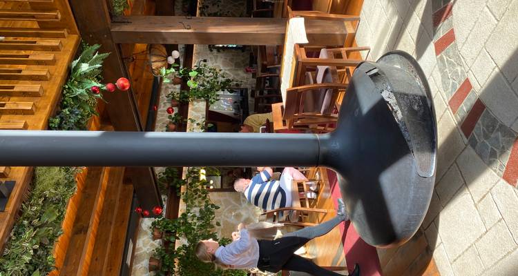 Interior de restaurante soleado con estufa de leña negra central y comensales en mesas de madera