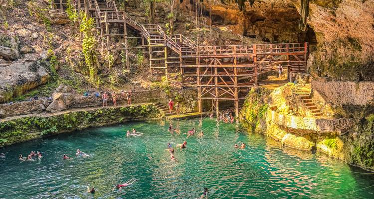 Drukke cenote met zwemmers in smaragdgroene water, houten terrassen en zonbeschenen grotwanden