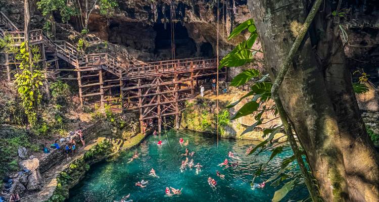 Verheven uitzicht op een cenote-grot met zwemmers en houten platforms in het zonlicht