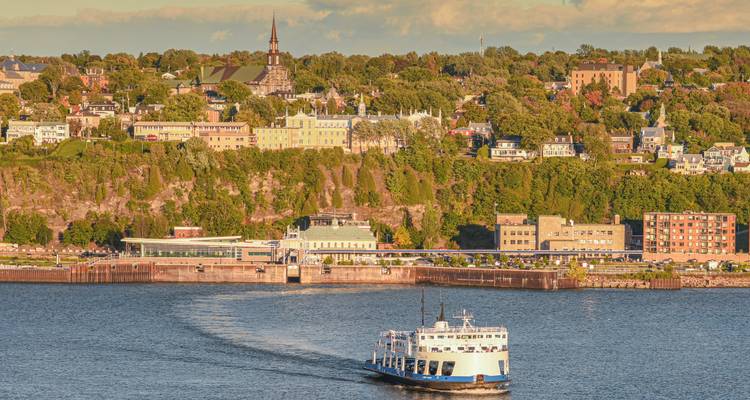 Un traversier pour passagers laissant un sillage sur un large fleuve avec les bâtiments de la Ville de Québec perchés sur les falaises baignés dans la lumière chaude du soir.