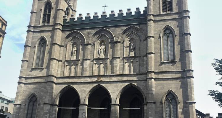Vue de face d'une basilique en pierre grise avec des tours pointues et des portes en arc sous un ciel dégagé.