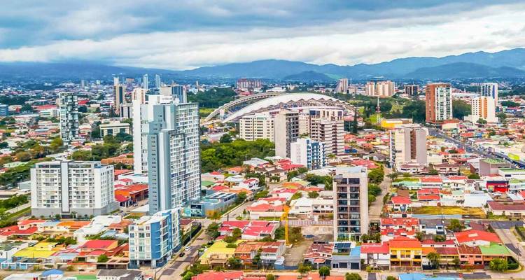 Panorámica aérea de una ciudad latinoamericana moderna con techos coloridos, edificios altos y montañas lejanas bajo cielos nublados