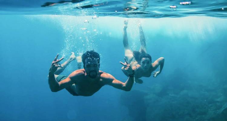 Deux plongeurs en apnée nagent sous l'eau dans une mer bleu clair, l'un d'eux faisant le signe de la paix.