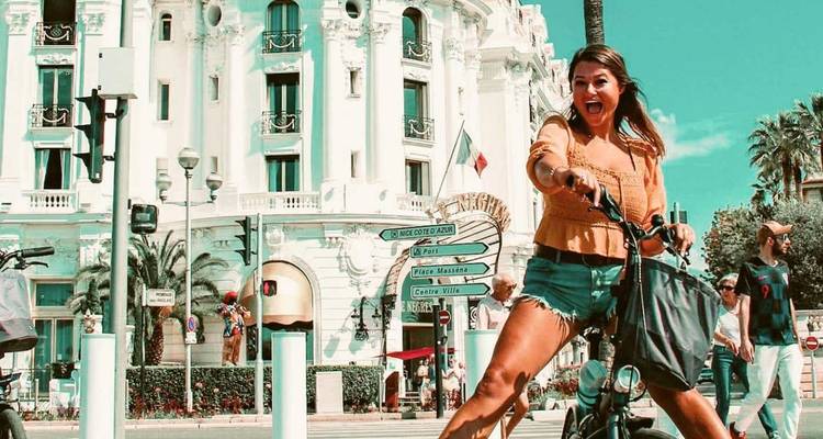 Femme enthousiaste sur une trottinette électrique passant devant l'orné Hôtel Negresco blanc et la promenade bordée de palmiers.
