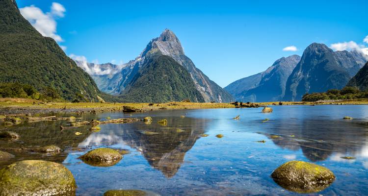 Le pic Mitre se reflétant dans les eaux calmes du Milford Sound, encadré par des montagnes luxuriantes et un ciel bleu.