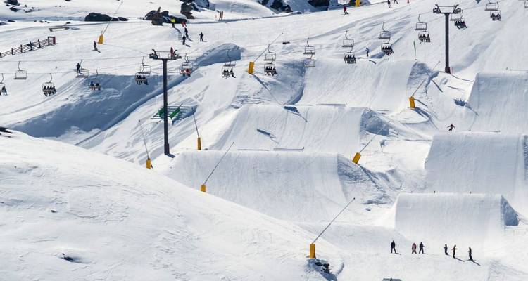 Parc alpin enneigé avec remontées mécaniques, tremplins et skieurs dispersés sur des pentes d'un blanc éclatant.