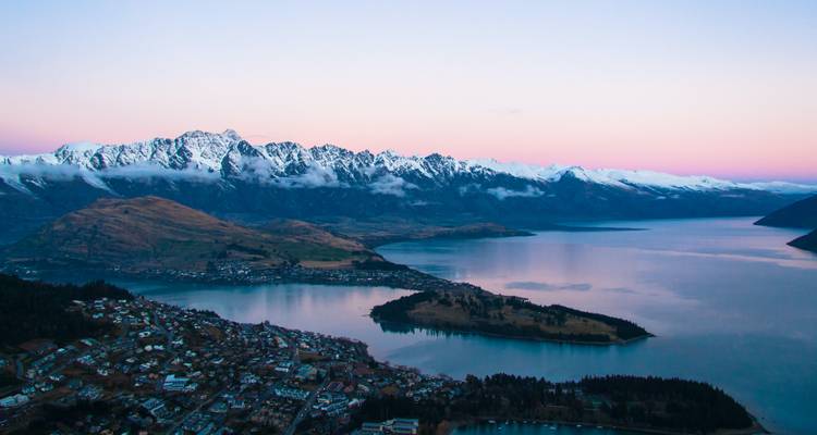 Vue crépusculaire en haute altitude surplombant une ville au bord d'un lac encadrée par des montagnes enneigées et des eaux calmes.