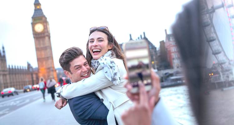 Energetic couple laughing and hugging on Westminster Bridge while a friend snaps a phone photo; Big Ben behind.