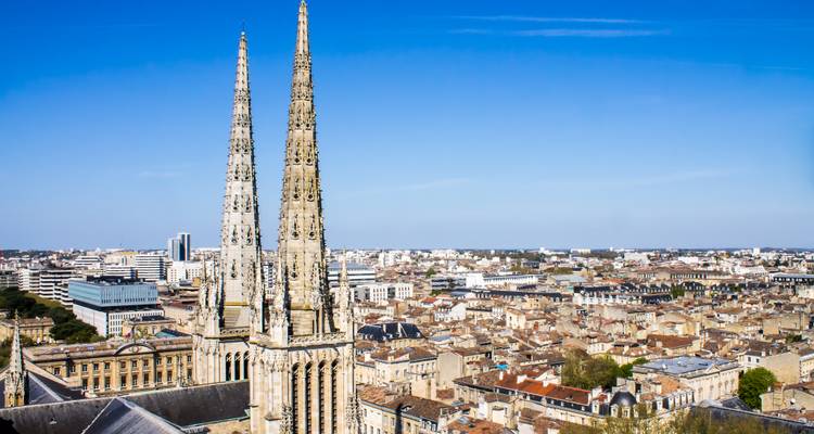Vue aérienne dégagée sur une ville européenne historique avec deux hautes flèches gothiques s'élevant au-dessus des toits en tuiles sous un ciel bleu.