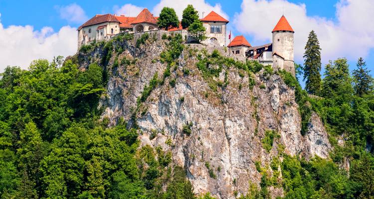 Le château de Bled, perché sur une falaise rocheuse entouré d'une forêt verdoyante et dominé par un ciel bleu et des nuages.