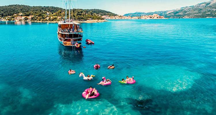 Un velero de madera se ancla en las aguas turquesas y vibrantes del Adriático mientras los viajeros flotan en anillos rosas cerca de ahí.