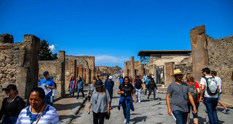Rue animée de la Pompéi antique avec de nombreux visiteurs marchant entre les ruines de pierre sous un ciel dégagé.