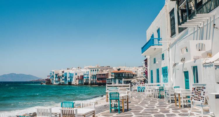 Des tables de café le long de la promenade pavée de la Petite Venise de Mykonos donnant sur les mers turquoise et les maisons colorées du front de mer.