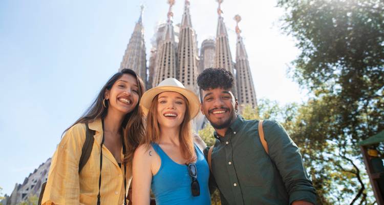 Tres amigos sonríen para una selfie frente a las torres imponentes de la Sagrada Família en un día soleado.