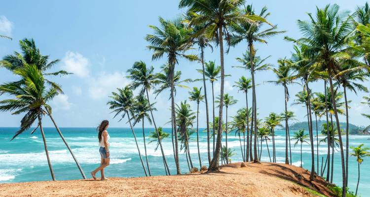 Une femme se tient sur un promontoire couvert de palmiers à terre rouge, surplombant les vagues turquoise de l'océan Indien.