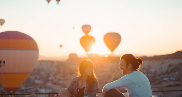 Un couple discutant sur une terrasse au coucher du soleil avec des montgolfières flottant sur le fond brumeux de la Cappadoce.