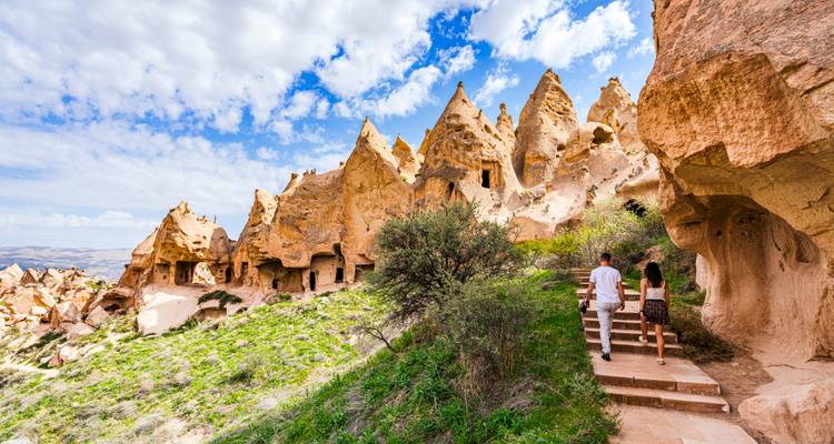 Un couple monte les marches de pierre parmi les formations rocheuses spectaculaires des cheminées de fée de la Cappadoce sous un ciel bleu éclatant avec des nuages épars.