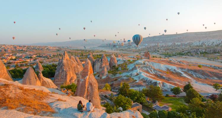 Les montgolfières dérivent au-dessus des vallées surréalistes et des pics de la Cappadoce au lever du soleil tandis qu'un couple est assis sur un rebord rocheux en admirant la vue.
