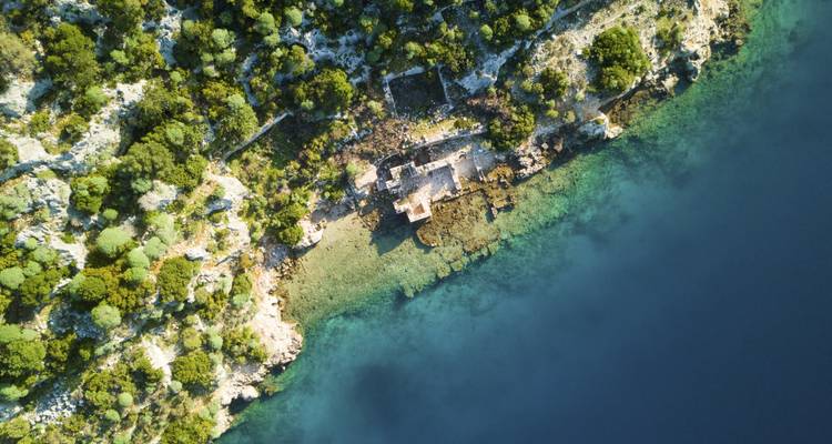 Vue aérienne d'un littoral turquoise avec des ruines de pierre anciennes nichées parmi une verdure luxuriante et une eau bleu clair.
