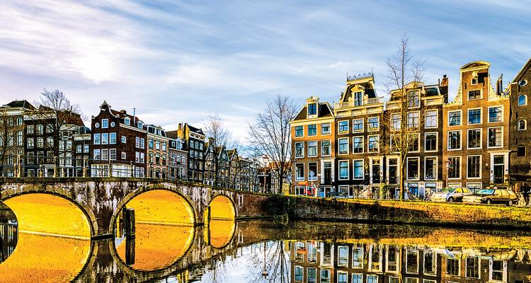 Pont en arc de pierre doré par la lumière dorée et maisons à pignon au bord du canal se reflétant parfaitement dans les eaux calmes d'Amsterdam à l'aube.