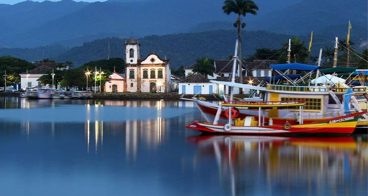 Abendliche Ansicht der Paraty-Uferfront mit Kolonialkirche, die sich im ruhigen Wasser vor einer Bergkulisse spiegelt.