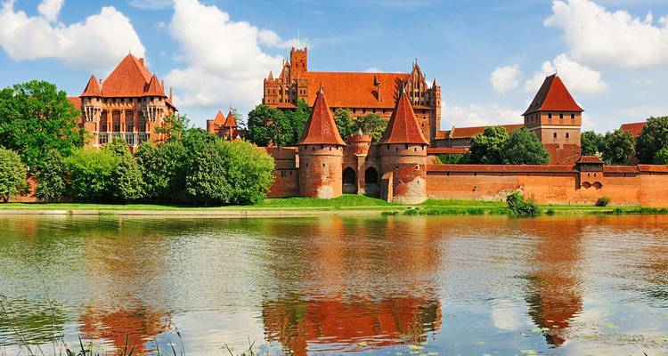 Le château de Malbork en brique rouge se reflétant dans une rivière calme sous un ciel bleu éclatant.