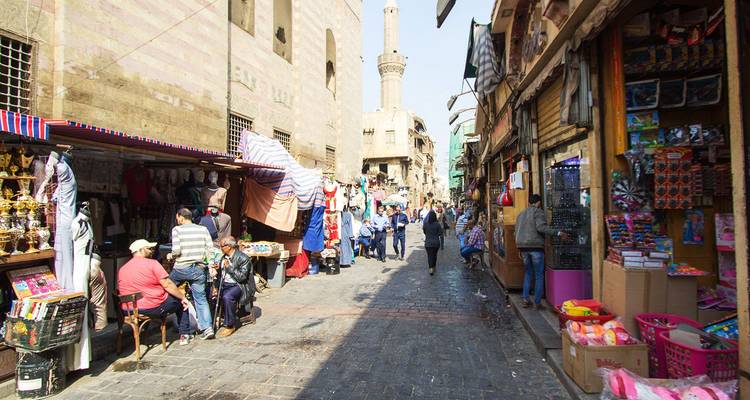 Lebendige Marktstraße im historischen Kairo mit Verkäufern, Einkäufern und einem entfernten Minarett.