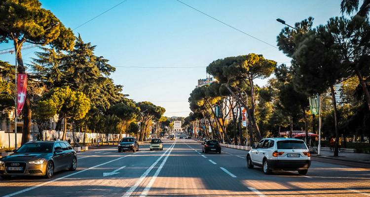 Brede, met bomen omzoomde boulevard waar auto's 's middags in de zon richting het centrum van Tirana rijden.