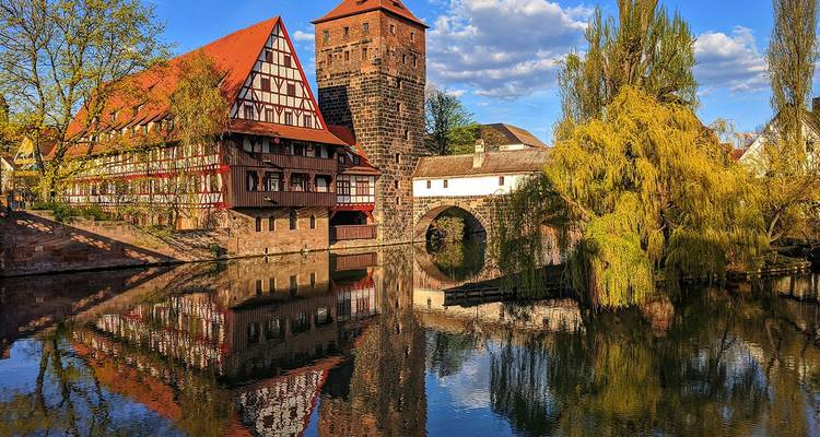 Fachwerkhaus, mittelalterlicher Turm und steinerne Brücke spiegeln sich in einem ruhigen Fluss in Nürnberg unter blauem Himmel wider.