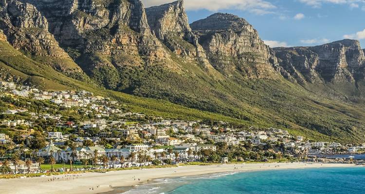 Vista aérea panorámica de la playa de Camps Bay enmarcada por aguas turquesas y la imponente cadena montañosa de los Doce Apóstoles salpicada de casas.
