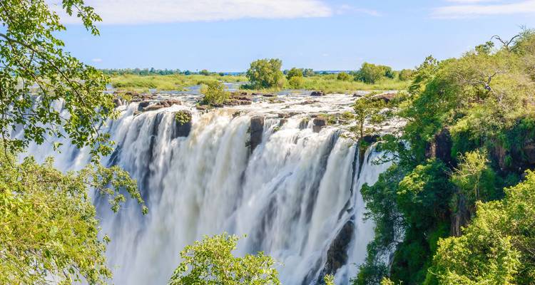Las poderosas Cataratas Victoria cayendo al desfiladero rodeado de vegetación bajo un cielo azul.