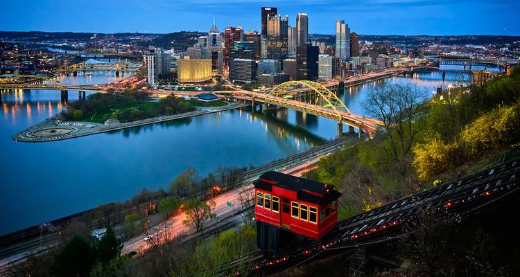 Vista dramática al atardecer sobre el horizonte de Pittsburgh y sus ríos con el teleférico rojo de Duquesne bajando.