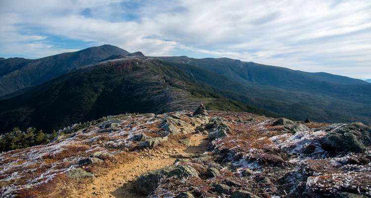 Sendero de cresta de las Montañas Rocosas con parches de escarcha temprana y picos expansivos bajo un cielo parcialmente nublado.