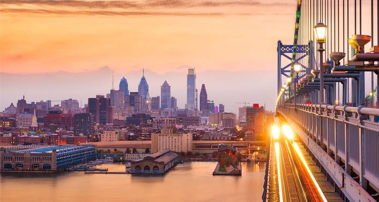 Skyline von Philadelphia, die bei Sonnenuntergang vom Ben Franklin Bridge aus leuchtet, mit Lichtspuren entlang der Gleise